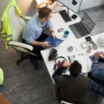 Three middle aged men including Caucasian foreman discussing construction project at office desk, reviewing blueprints and digital plans on laptop, safety vest and helmet visible nearby