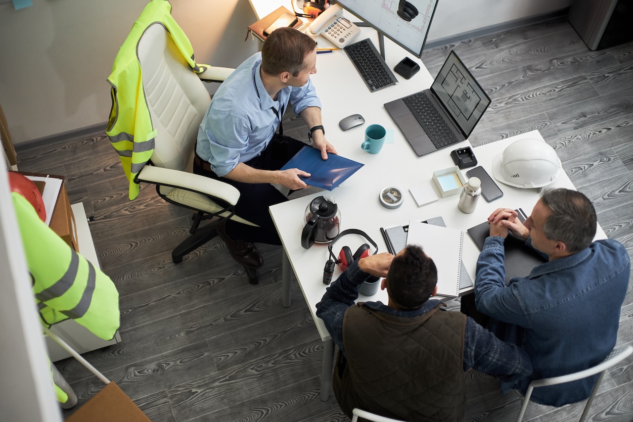 Three middle aged men including Caucasian foreman discussing construction project at office desk, reviewing blueprints and digital plans on laptop, safety vest and helmet visible nearby